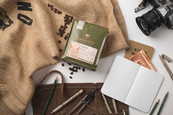 Adventure themed flatlay image with a burlap coffee bag, notebooks and pencils, a camera, and a bag of Canyon Coffee Roasters coffee