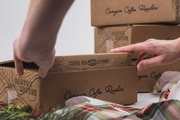 Person opening a cardboard box labeled 'Canyon Coffee Roasters' with more boxes stacked in the background.